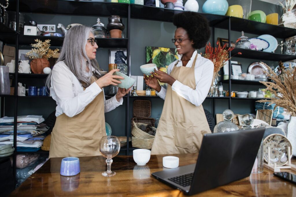Two women in aprons holding ceramic bowls at a shop counter, with shelves of gifts behind them