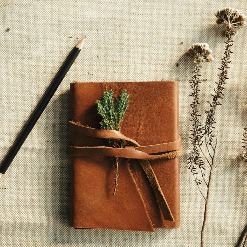 Leather-bound journal tied with a strap, beside a pencil and dried flowers on linen
